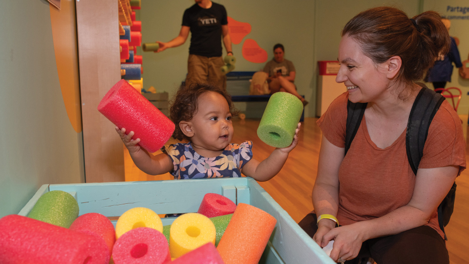 A toddler, accompanied by a smiling adult, holds up several colourful foam shapes.
