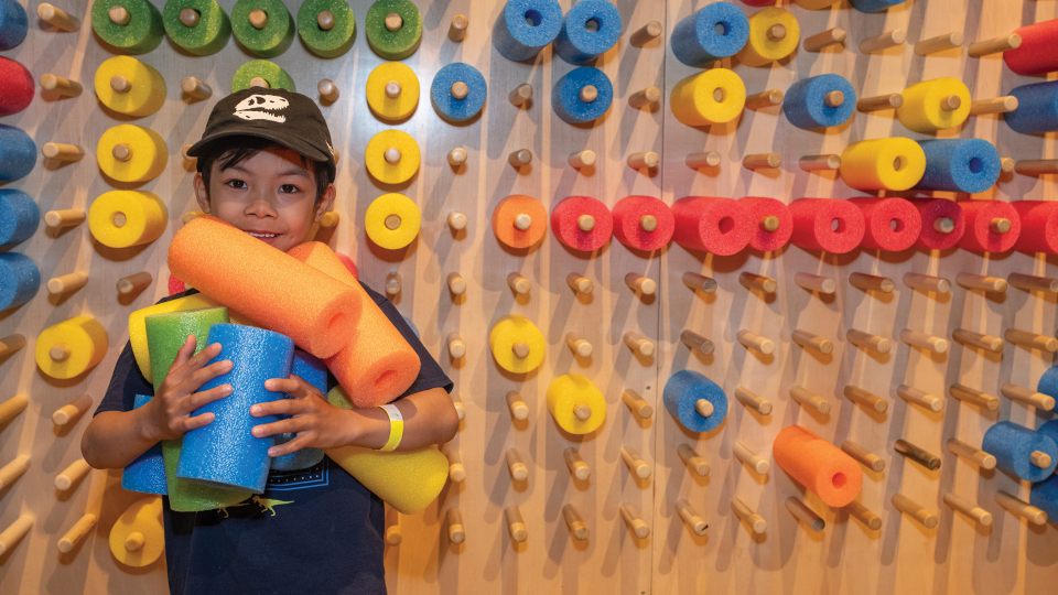 A young child, holding an armful of colourful foam noodle segments, stands in front of a wall covered in "noodle" pegs.
