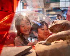 Two smiling children look through a clear dome display at a prehistoric reptile model in a museum exhibit.