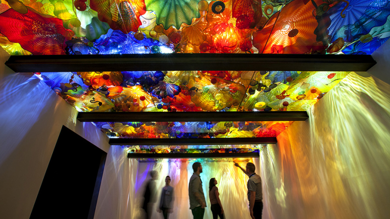 Visitors in the exhibition with glass sculptures displayed above a back lit glass ceiling. 