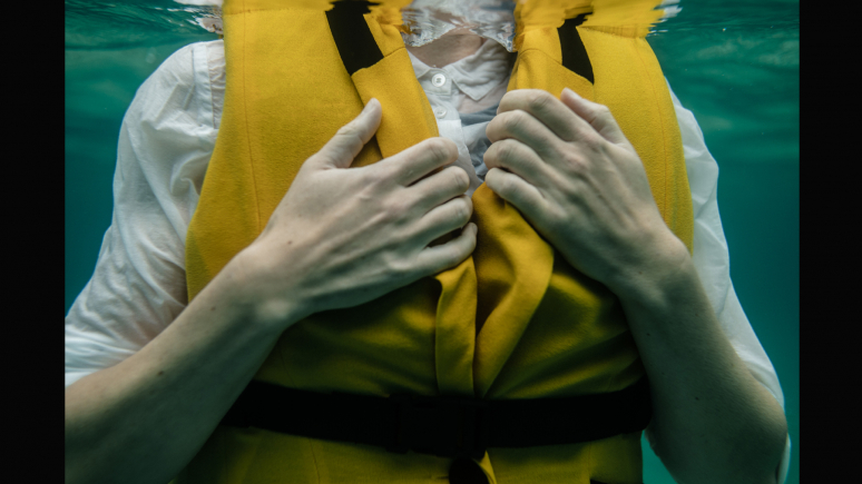 Image taken underwater of a woman's torso fited in a white shirt and yellow lifejacket.