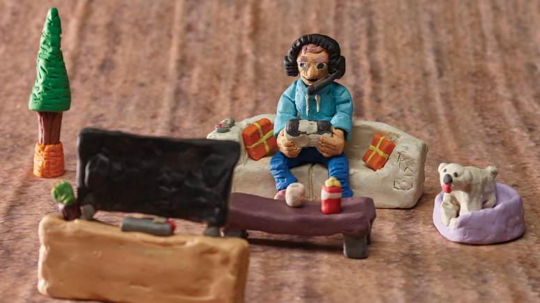 Detail of a diorama showing a person wearing headphones on a couch, playing a video game while looking at a television screen.