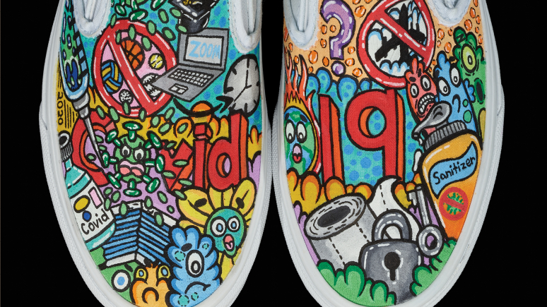 Image detail of a pair of white, slip-on sneakers with the tops painted in a colourful mosaic of COVID images and symbols.