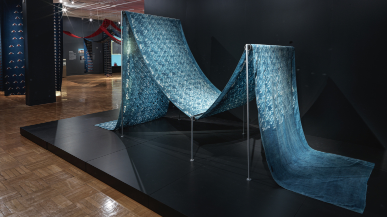 View of a length of dyed blue cotton fabric with mirror embellishments arranged on a frame to resemble fabric on a weaving loom in the exhibition Swapnaa Tamhane: Mobile Palace, at ROM.