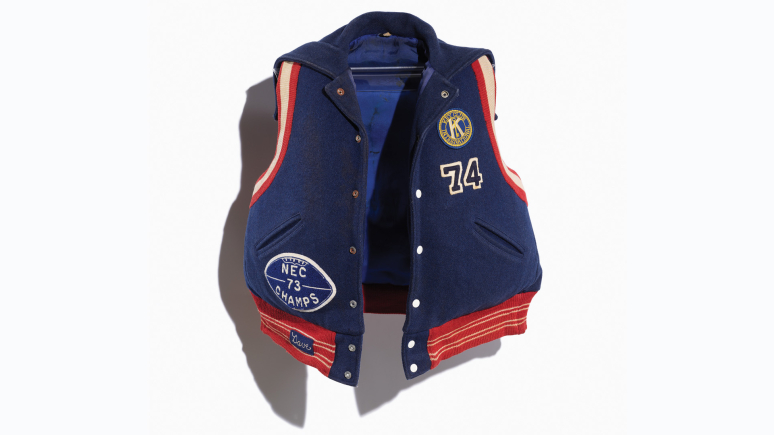 Image of a repurposed life jacket covered in a navy varsity jacket with orange and red trim and school letters and team patches/numbers. Image taken on a white background.