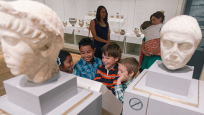 Four young children talk to each other while smiling in front of two ancient Roman busts.