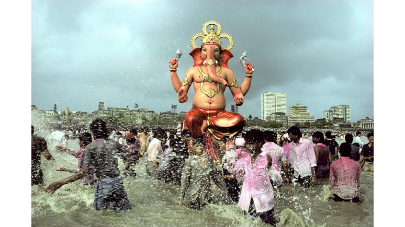 People walking through water.  In the center and high above the crowd is a respected figure in seated position with four arms/hands. The face is an elephant.