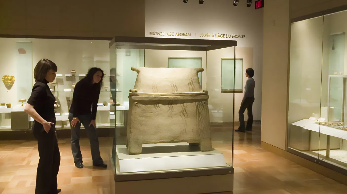 Two people examine a sarcophagus in a gallery, one leans forward to get a closer look at the details.