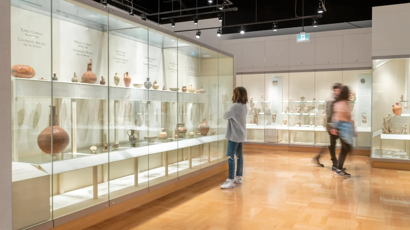 Three people walk around a gallery filled with ancient pottery in cases.