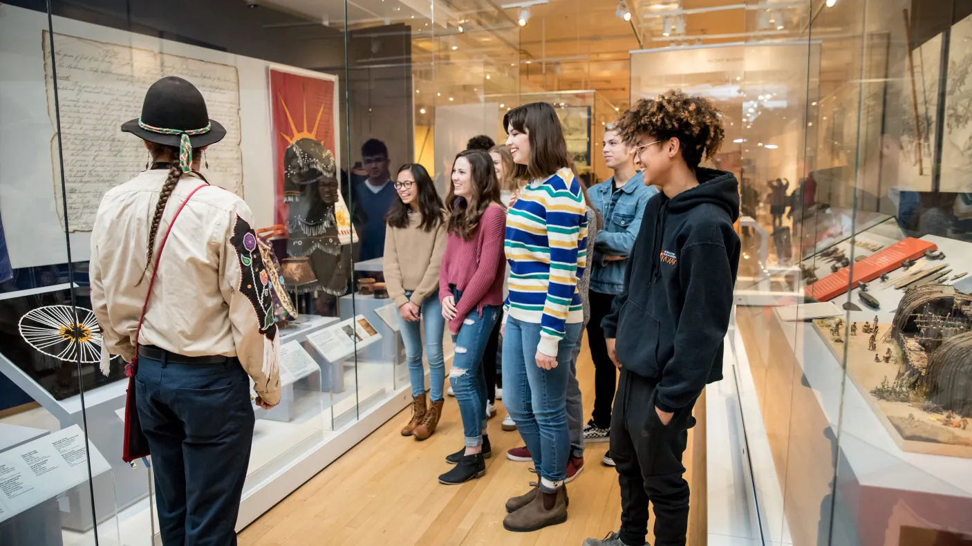 A museum educator in traditional Indigenous dress with braids and a black hat guides a group of diverse students through a history exhibit with glass display cases containing artifacts and documents.