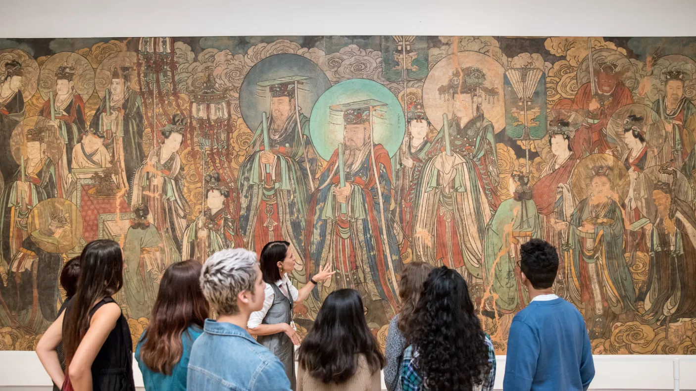 A group of students listens to a guide in front of a large traditional East Asian mural in a museum.
