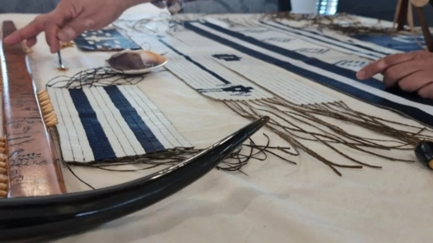 Close-up of hands working on a traditional wampum belt weaving with beads, threads, and tools laid out on a table.