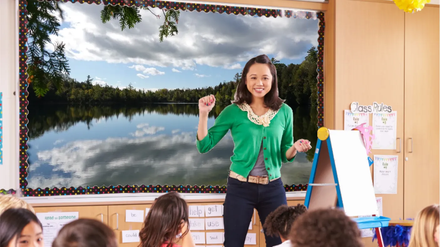 A teacher in a green cardigan stands in front of a smartboard at the front of a class. The board displays an image of a glassy lake.