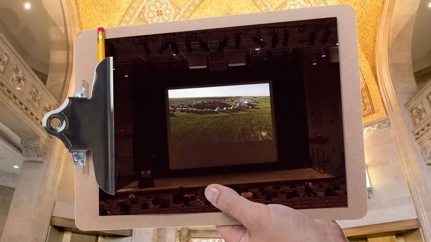 A hand holds a clipboard in front of a large gallery with a gold mosaic ceiling. The clipboard displays a picture of students in a darkened theatre watching a presentation.