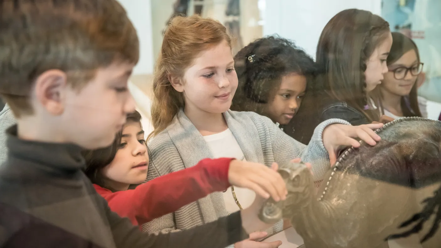 Several young students in a diverse class touching a bronze statue of a maiasaur.