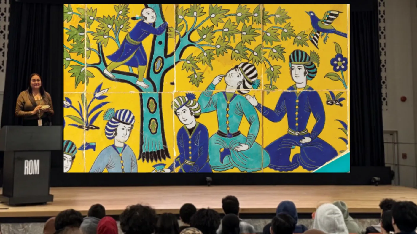A woman stands behind a podium on a stage. The screen behind her displays an image of a colourful Islamic tile depicting several people at a picnic.