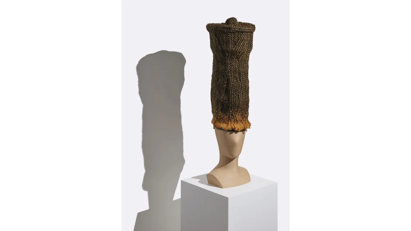 Tall cylindrical woven basket with a flared rim displayed on a white pedestal casting a shadow on the wall behind.