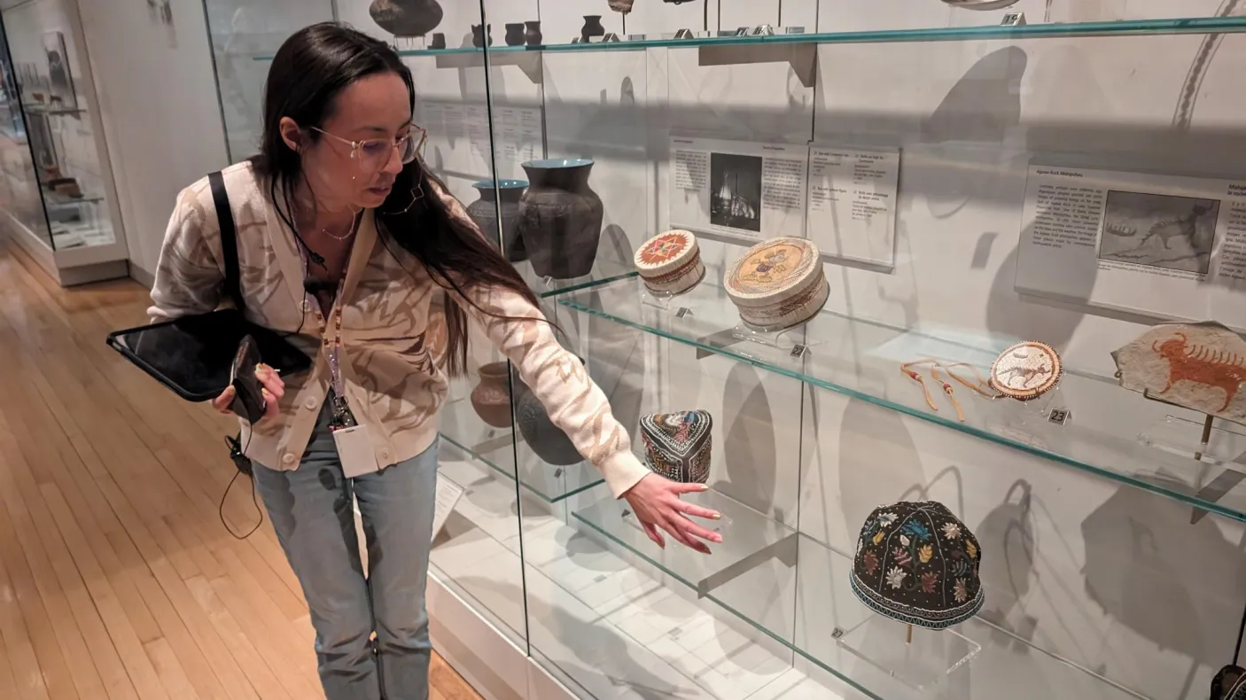 An Indigenous woman with long, dark hair and glasses stands by a museum case filled with belongings showing beadwork, quillwork, and other artistic traditions.