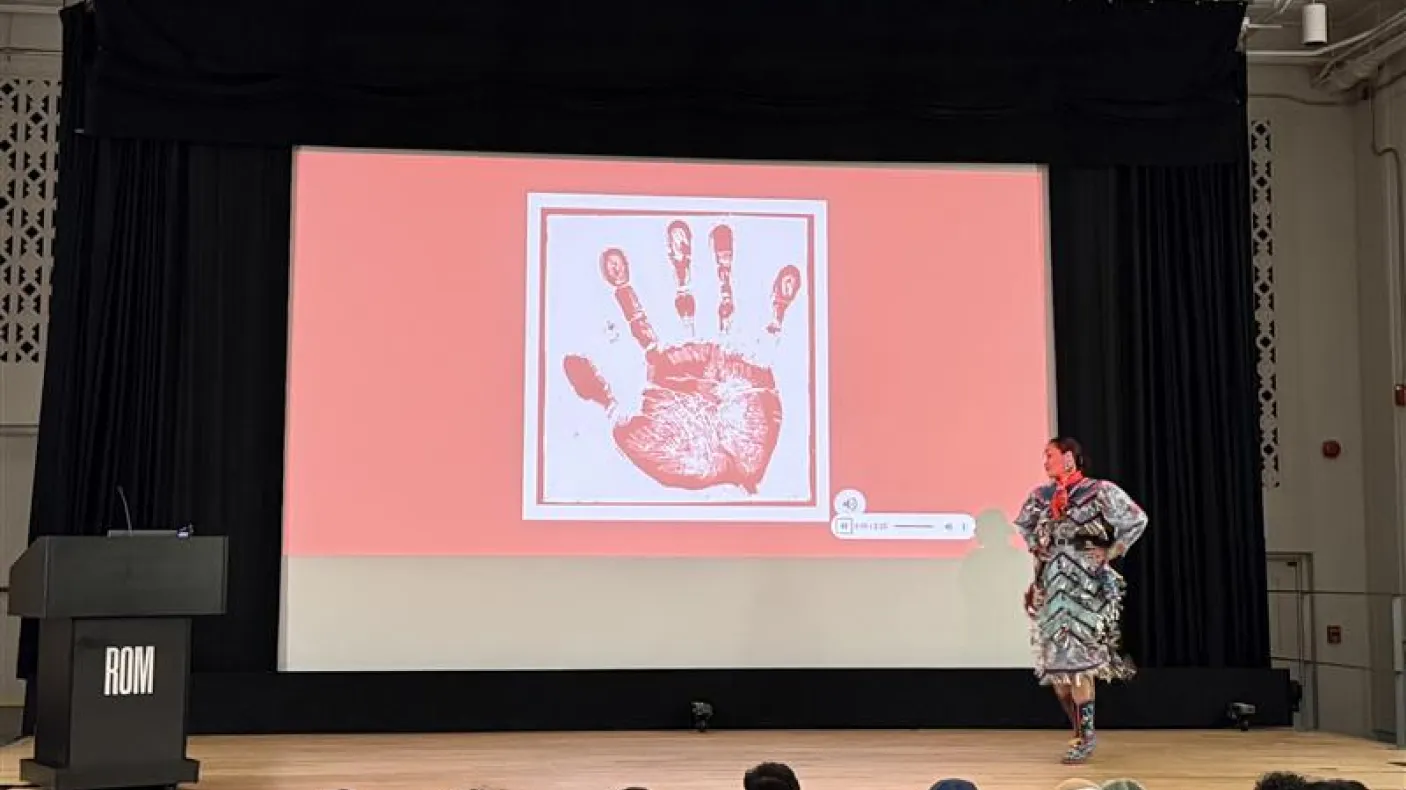 An Indigenous woman wearing a jingle dress stands on a stage in a theatre. The screen behind her shows a white handprint on a red screen.