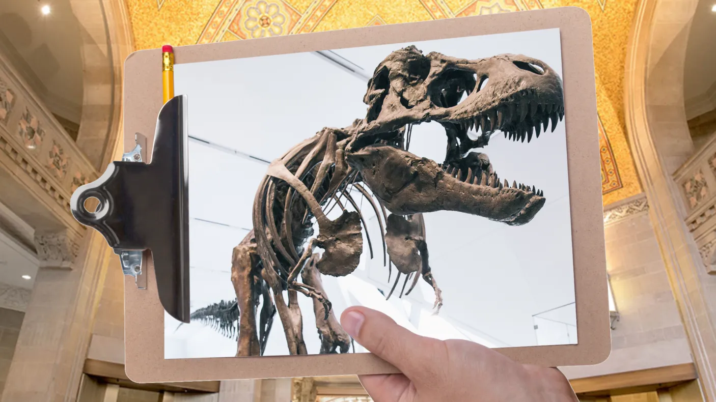 A hand holds a clipboard in front of a gallery with a mosaic ceiling. The clipboard is holding a picture of a Tyrannosaurus rex skeleton