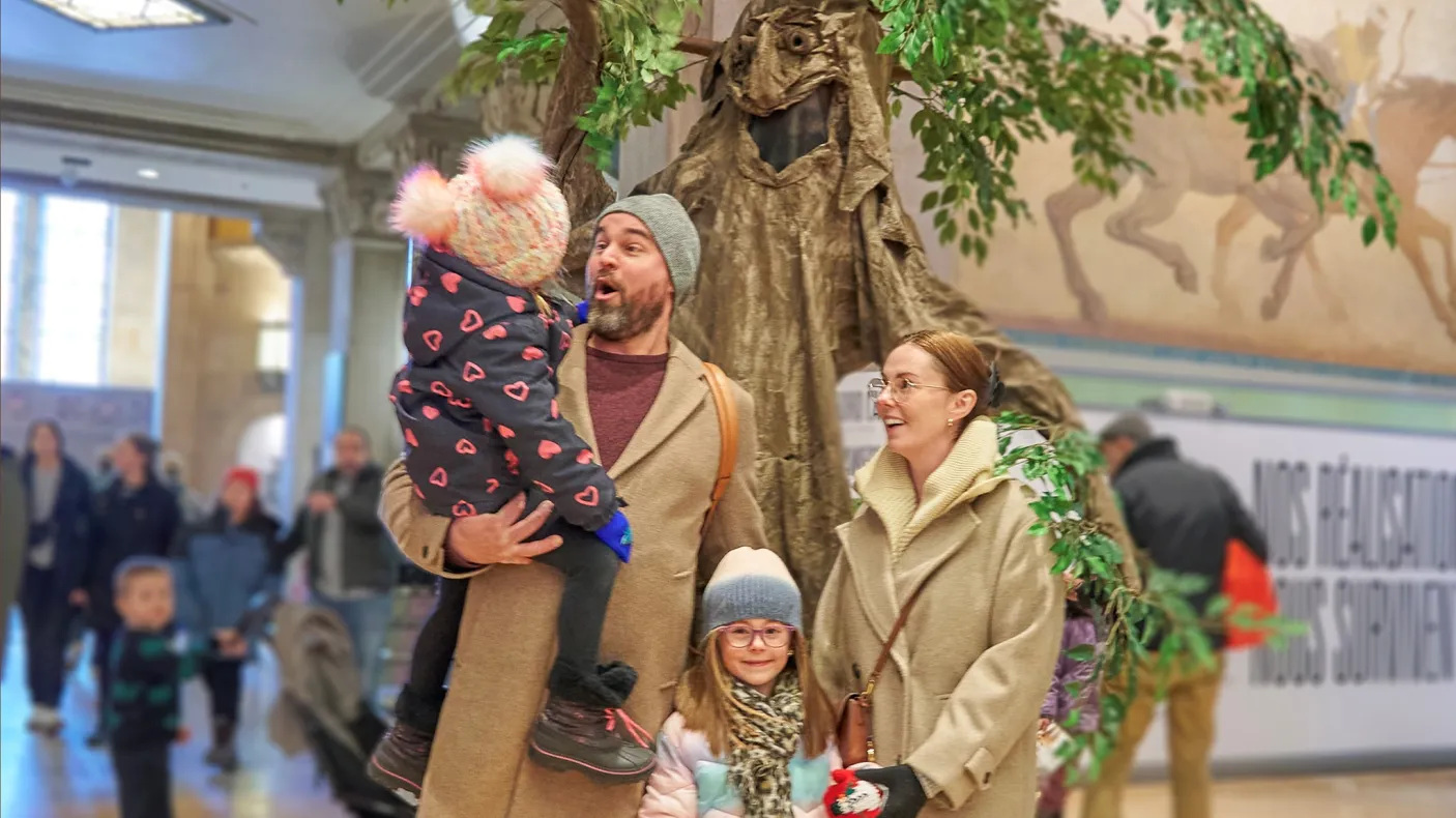 Two adults and two children stand indoors near a large tree-like display; one adult holds a bundled-up toddler while the others smile, with people walking in the background.