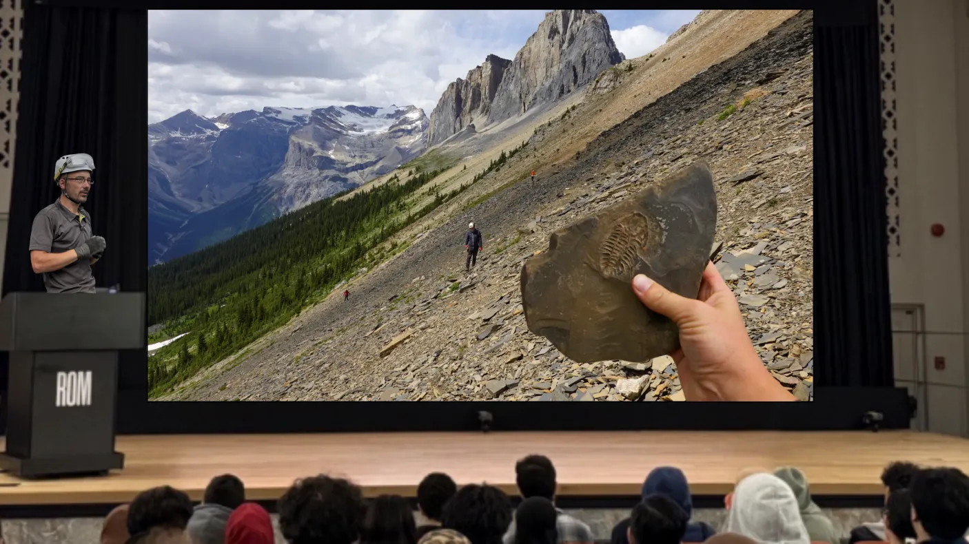 A man wearing a hard hat with goggles stands behind a podium on a stage. A screen next to him depicts a shale-covered hillside with mountains in the background. A lone figure walks across the hillside. A hand holds up a fossil in the foreground.