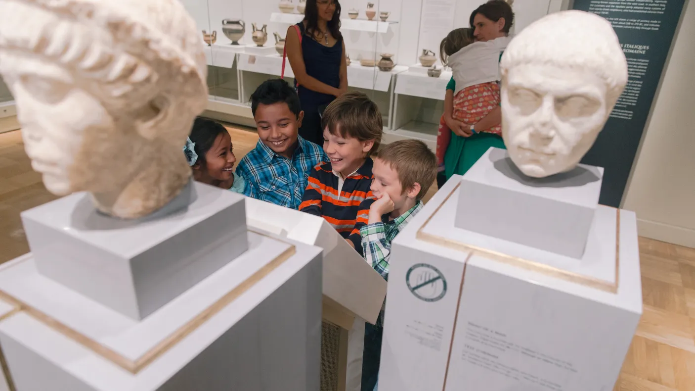 Two adults watch a smiling group of children in front of a group of marble busts