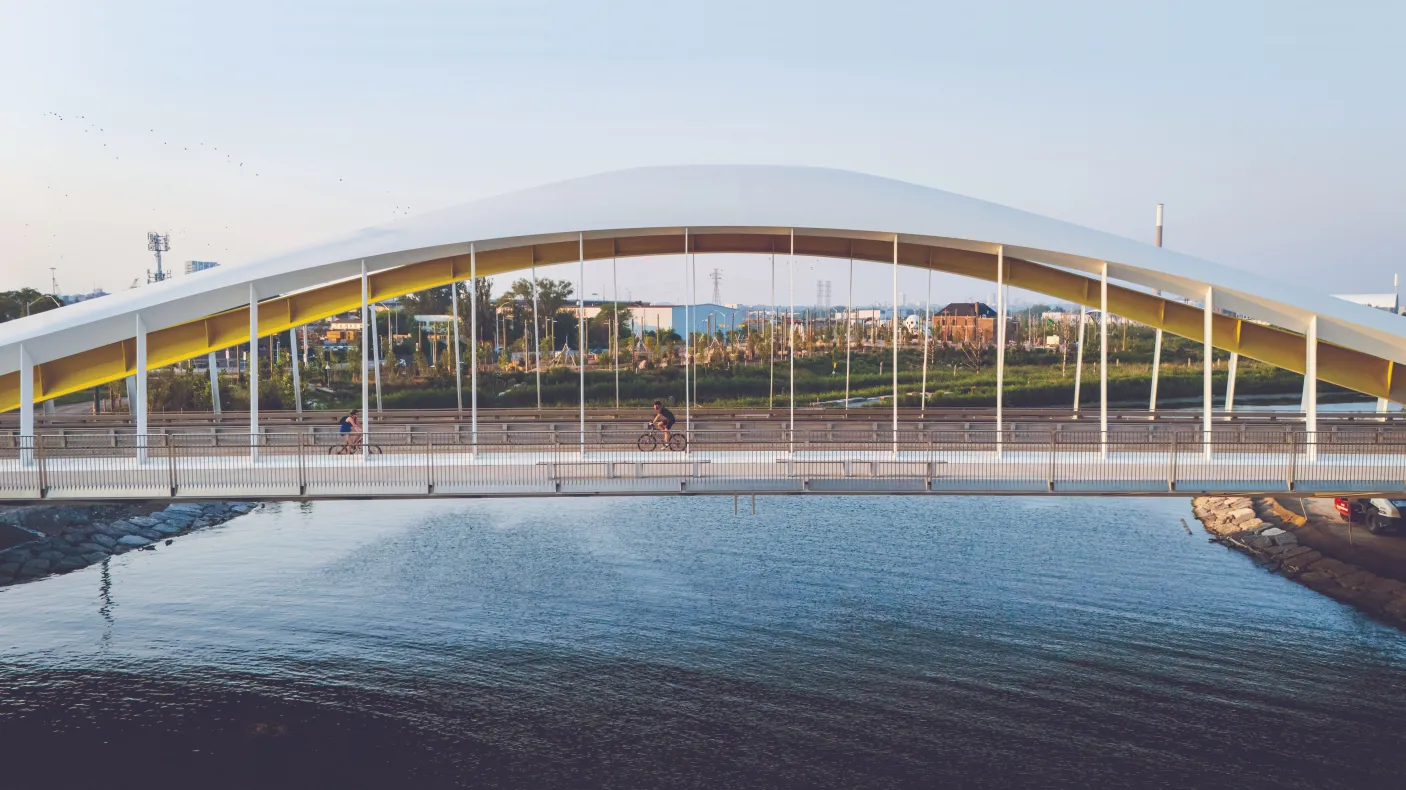 Wide view of a modern arched pedestrian bridge spanning a body of water, with cyclists crossing beneath a curved white canopy supported by vertical cables, set against a city landscape and pale blue sky in warm evening light.