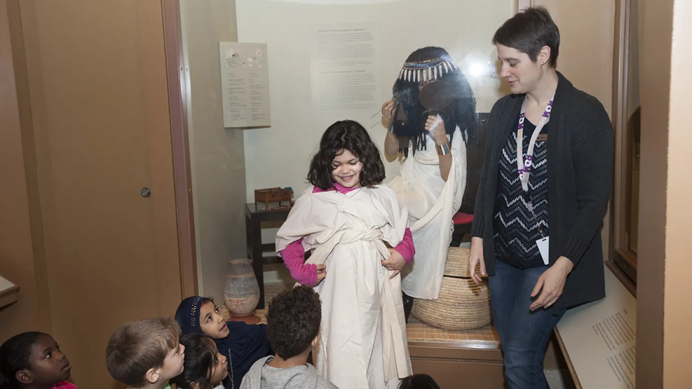A museum teacher dresses a smiling child in an Egyptian dress as classmates watch.