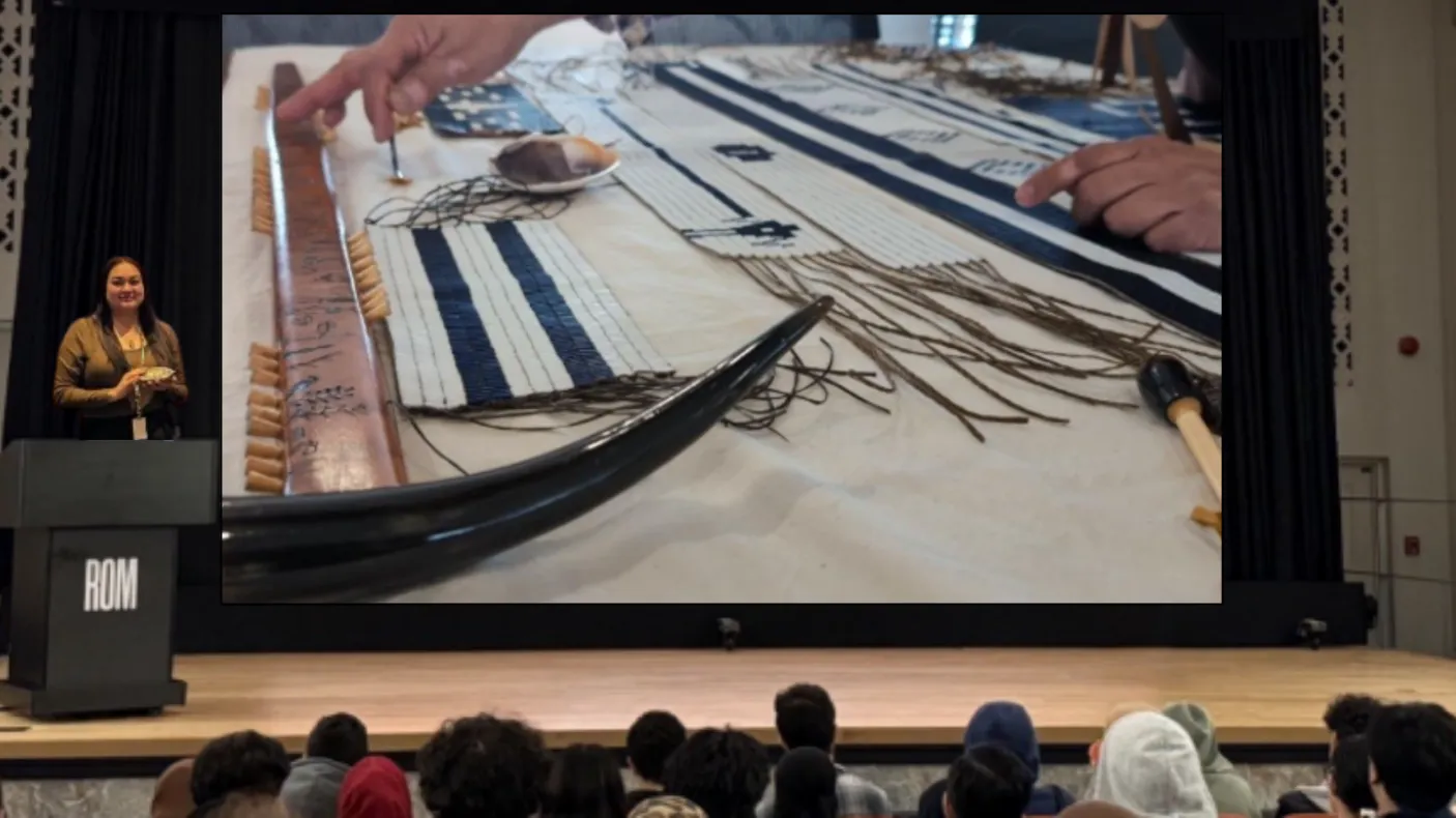 An Indigenous woman stands at a podium on a stage in front of a group of young people, gesturing to wampum belts and other belongings displayed on a screen.