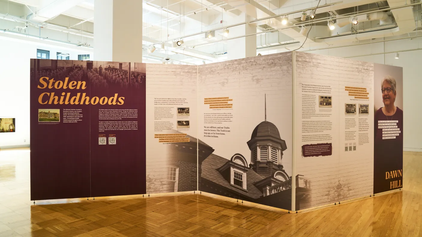 Large exhibition installation in a bright museum gallery featuring three angled display walls. The left panel, titled Stolen Childhoods, includes historic photographs and text about residential schools. The central panels display historical information, archival images, and a large black-and-white photograph of a school building. The right panel features a portrait and additional interpretive text. The exhibition stands on a polished parquet wood floor beneath high white ceilings with exposed lighting and d