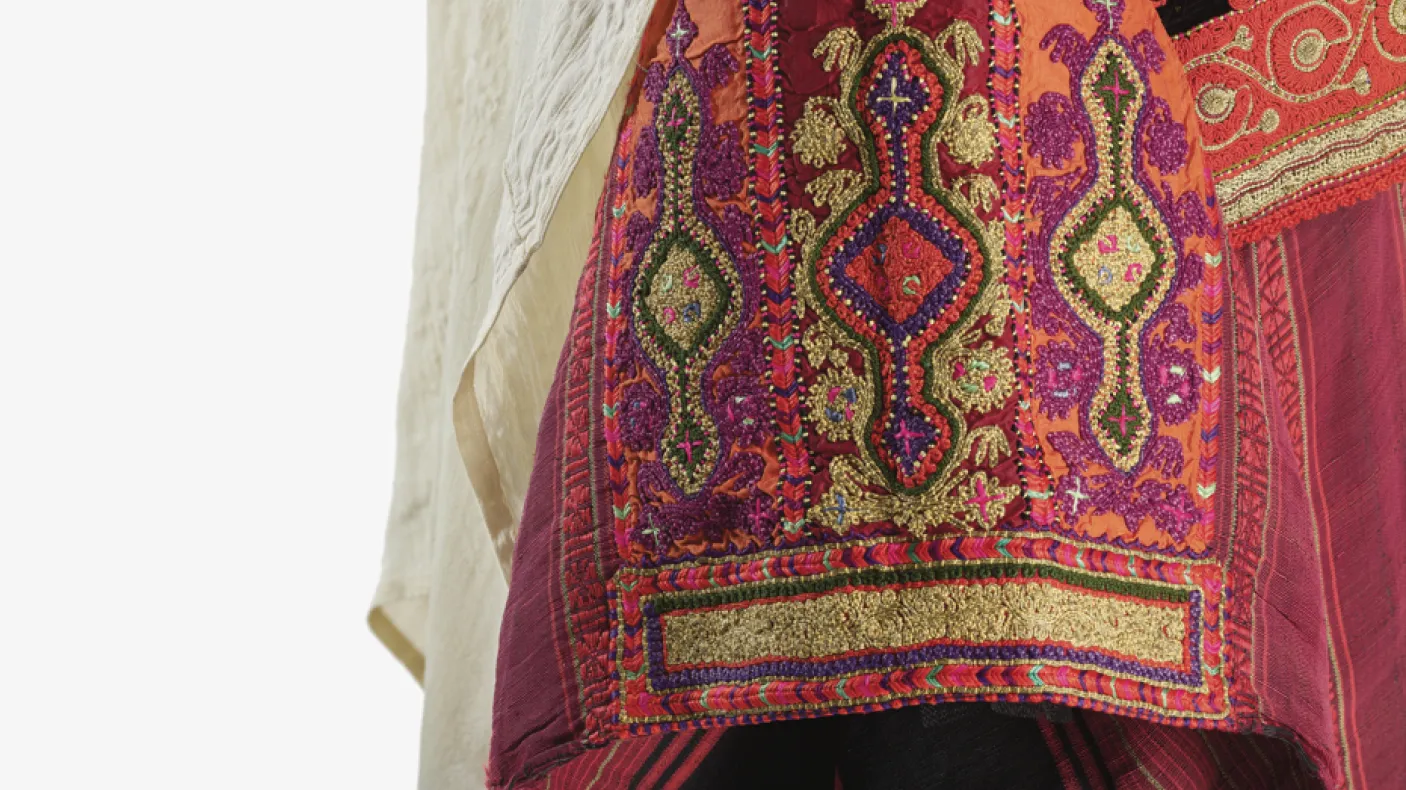 Close-up detail of a traditional garment featuring intricate red, orange, gold, and purple embroidery with geometric and floral patterns. A light-colored draped fabric falls alongside the richly textured textile, highlighting the craftsmanship and decorative stitching.