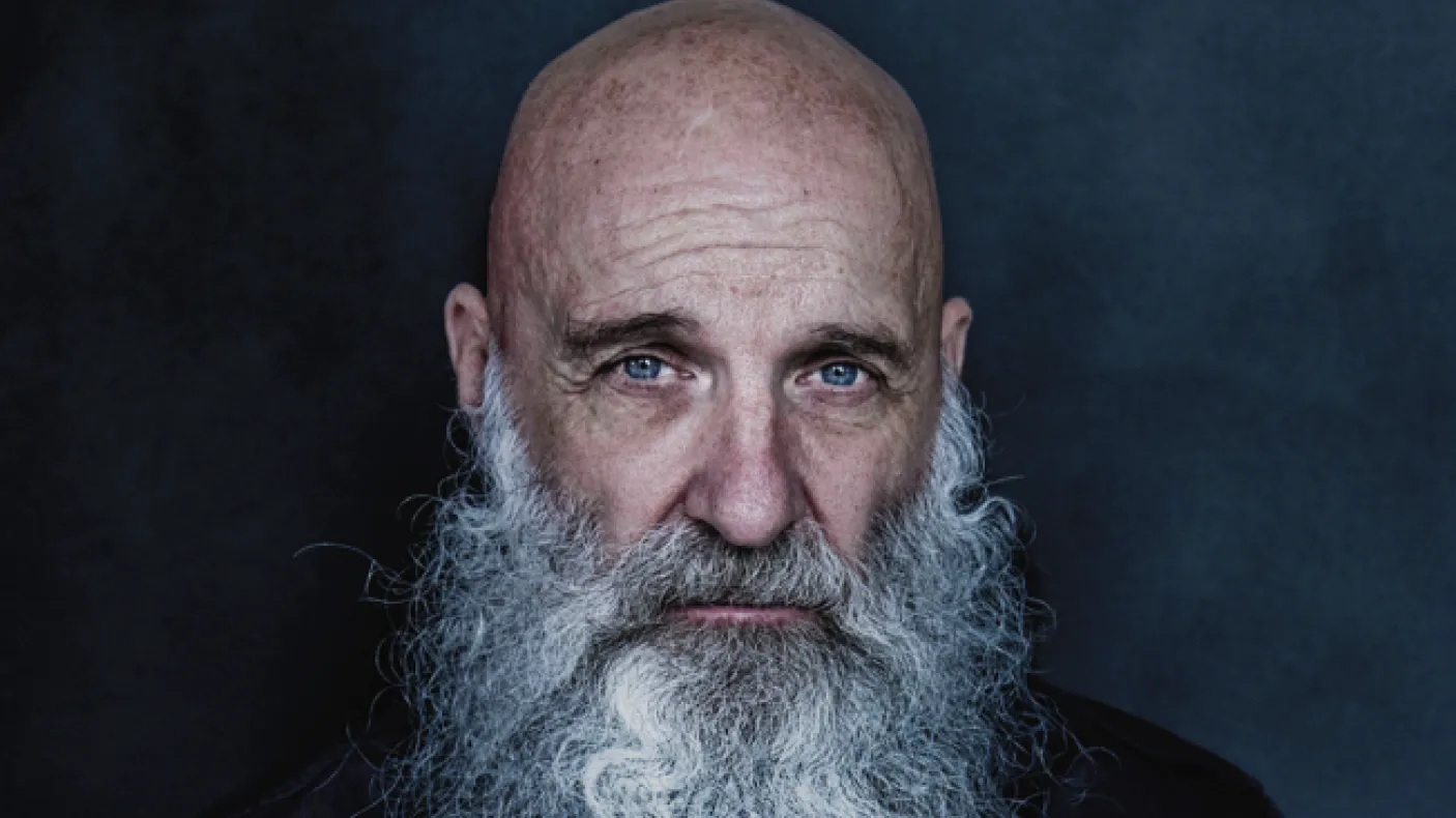 Studio portrait of a bald individual with a long white beard, wearing a dark button-up shirt against a dark blue background.