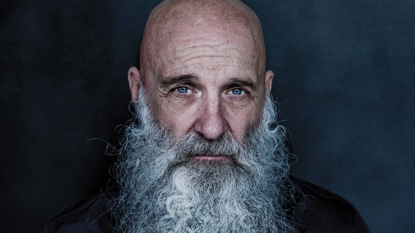 Studio portrait of a bald individual with a long white beard, wearing a dark button-up shirt against a dark blue background.