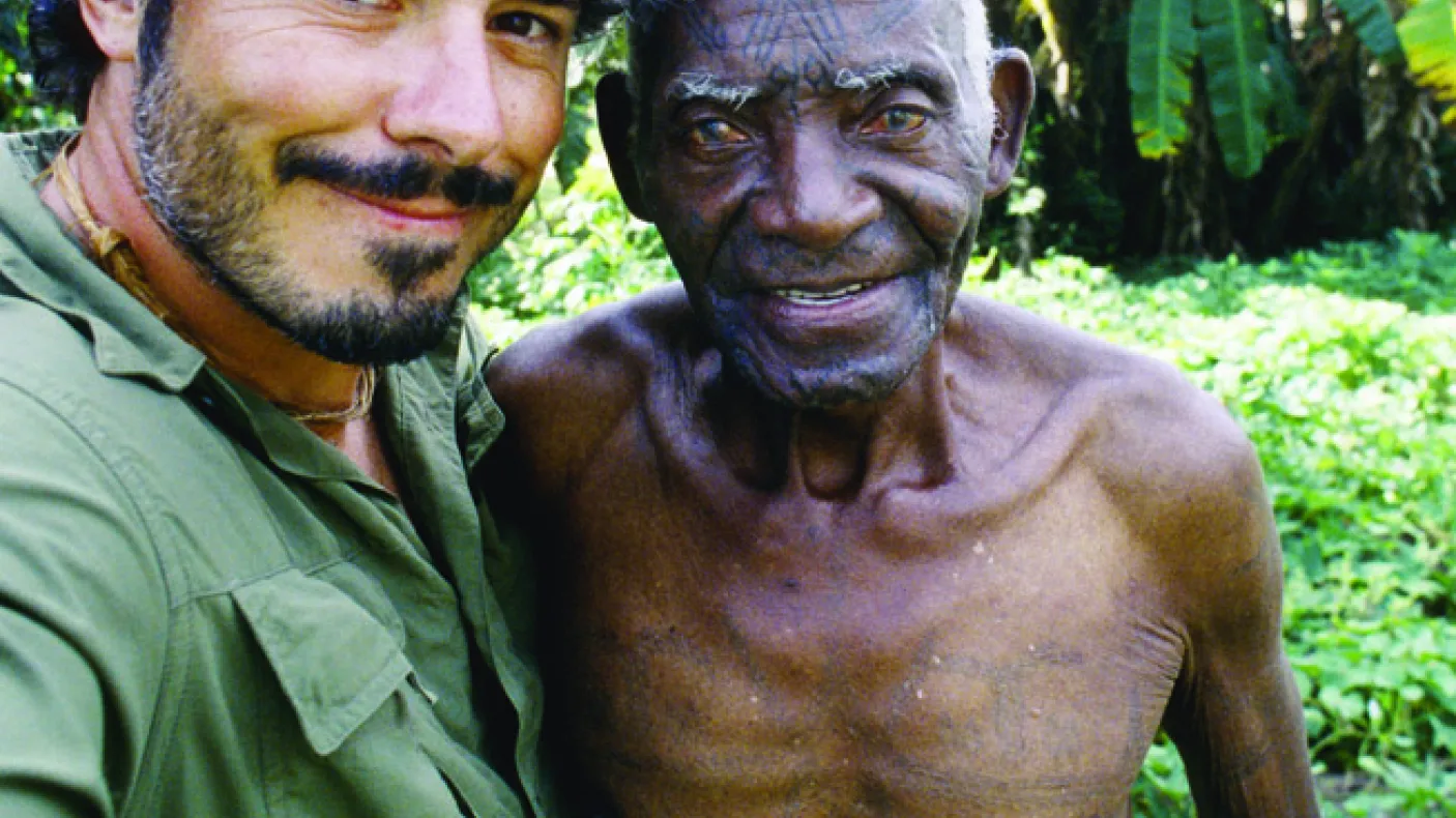 A man in a green shirt and blue bandana taking a selfie with a Makonde tattoo master
