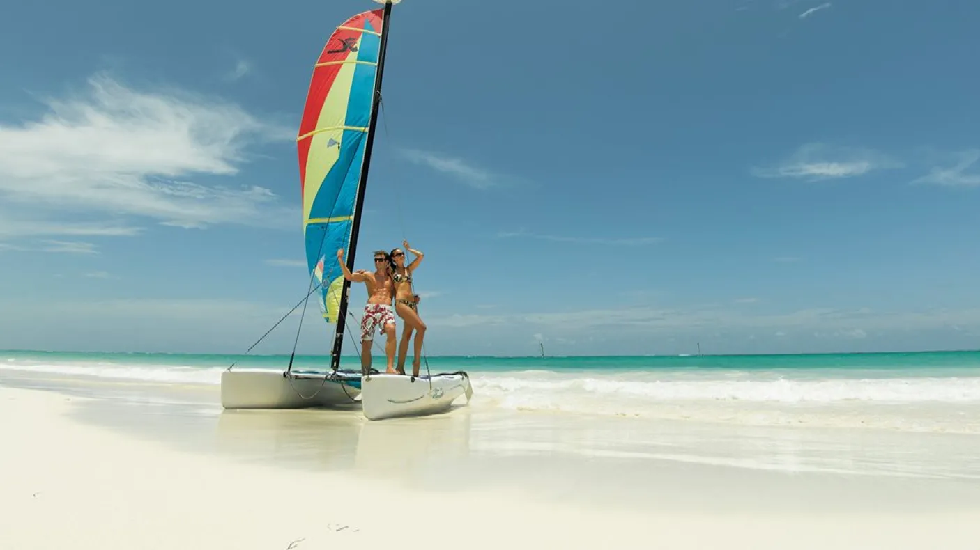 A man & a woman in bathing suits pose on a small catamaran on a sun-lit, white sand beach

