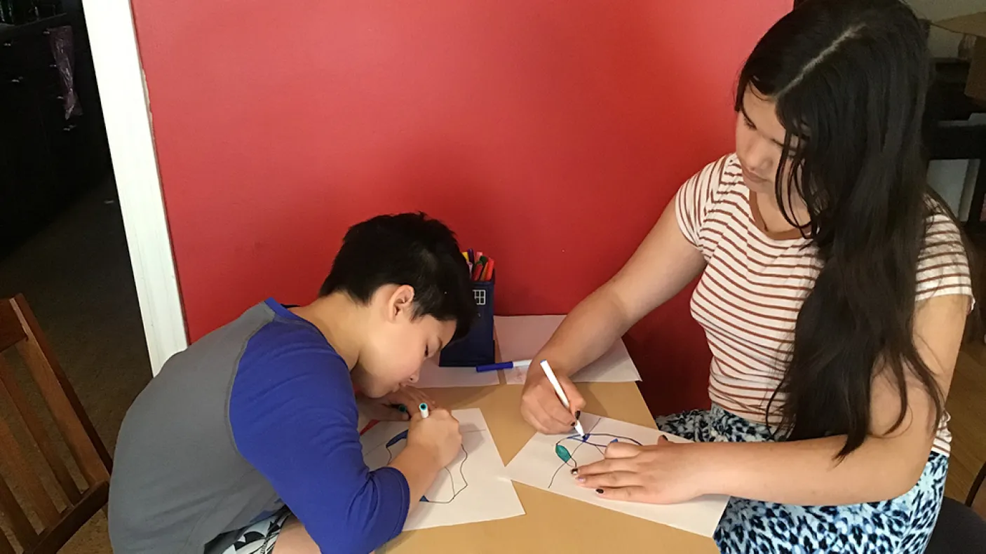 wo girls, one with short dark hair and the other with long dark hair, sit at a table and colour iceberg drawings with markers.
