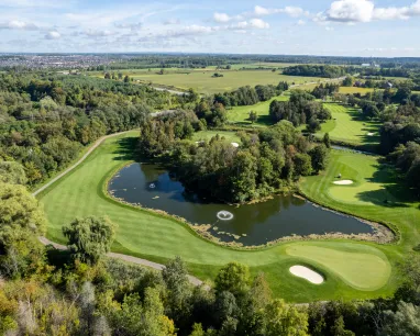 Aerial view of a golf course with a water hazard, sand traps, and surrounding trees under a blue sky.
