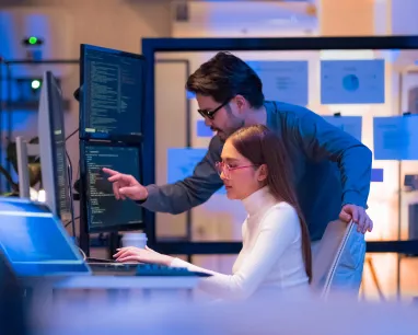A man stands beside a seated woman working on a computer, both looking at a monitor with code in an office setting.