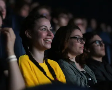 People seated in a dark theater watching a screen, with a person in a bright yellow shirt in the foreground.