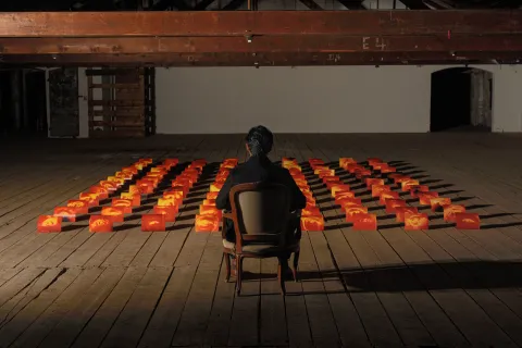 A person sits alone in a wooden chair facing an art installation of glowing orange and red blocks arranged in a grid, each block featuring a human eye, set in a dimly lit industrial-style room with exposed beams and wooden flooring.