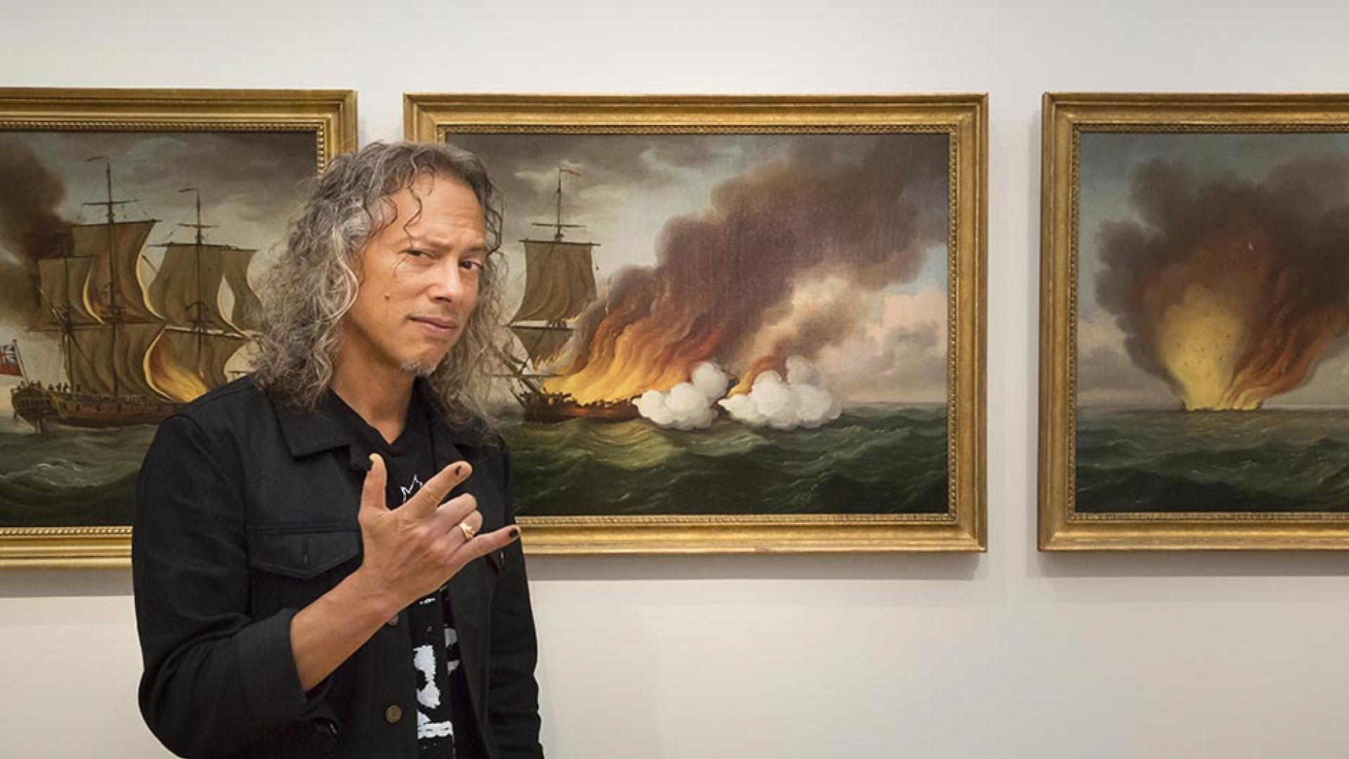 Kirk Hammett standing in front of three paintings of burning ships.