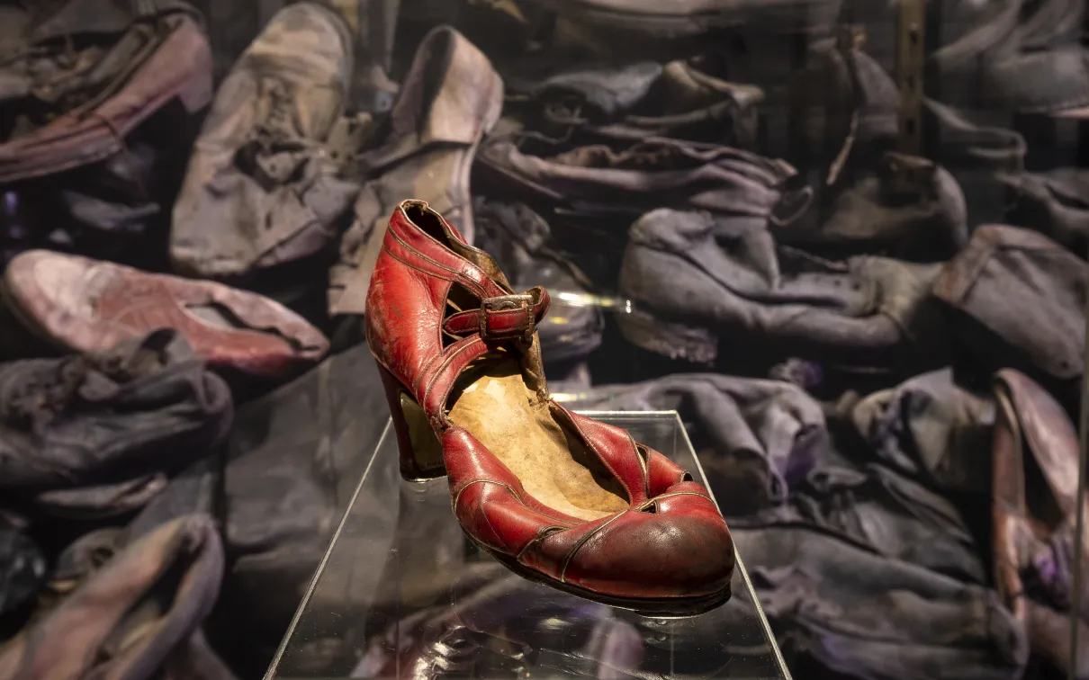 Une chaussure à talon haut rouge, abîmée et usée, présentée devant une pile de centaines d'autres chaussures.