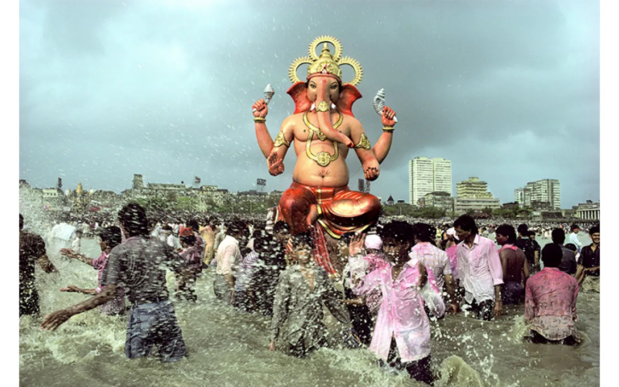 People walking through water.  In the center and high above the crowd is a respected figure in seated position with four arms/hands. The face is an elephant.