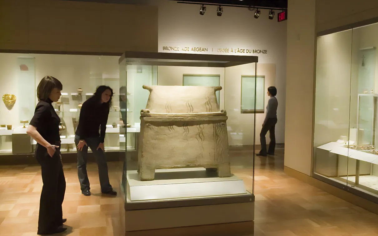 Two people examine a sarcophagus in a gallery, one leans forward to get a closer look at the details.