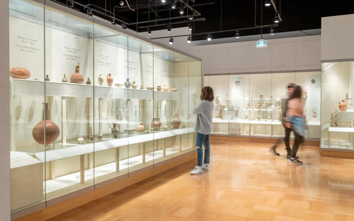 Three people walk around a gallery filled with ancient pottery in cases.