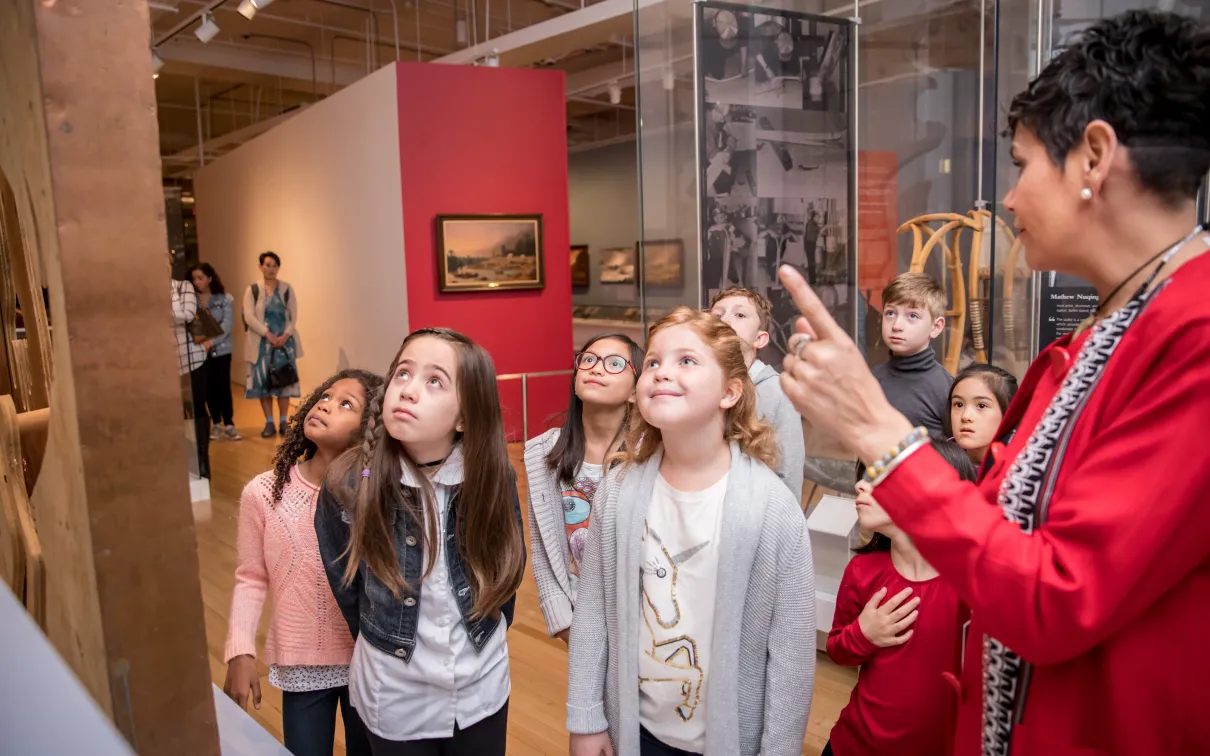 A group of children listens to a guide explaining an exhibit.