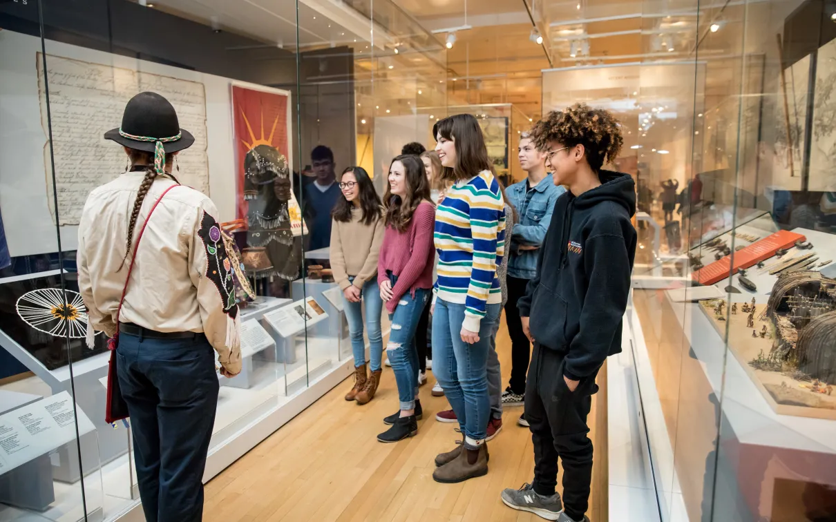 A museum educator in traditional Indigenous dress with braids and a black hat guides a group of diverse students through a history exhibit with glass display cases containing artifacts and documents.