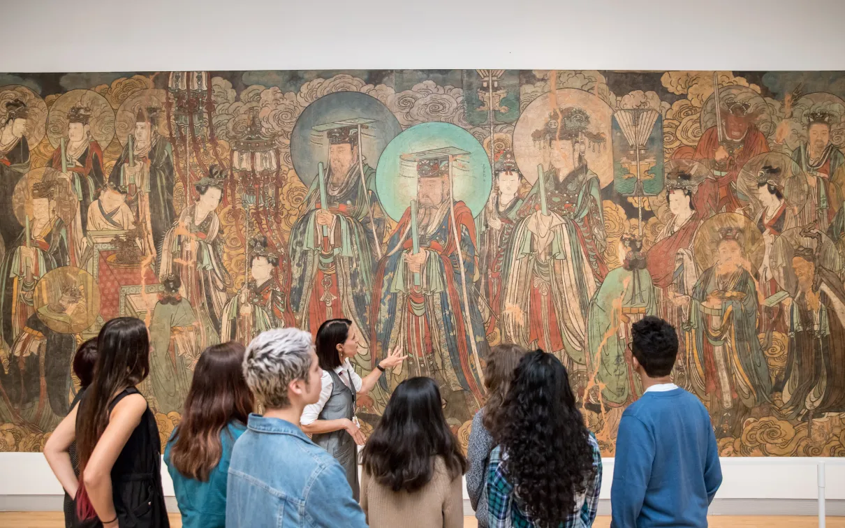 A group of students listens to a guide in front of a large traditional East Asian mural in a museum.
