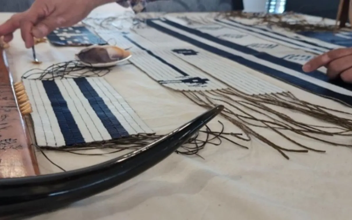 Close-up of hands working on a traditional wampum belt weaving with beads, threads, and tools laid out on a table.